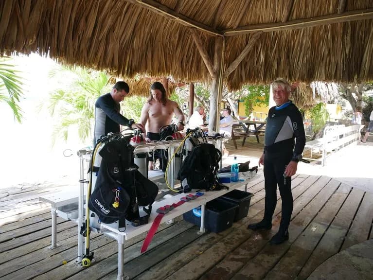 Three men in wet suits standing in front of a hut near the water's edge, ready for an aquatic adventure.